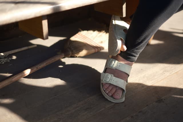 A person wearing sandals and black pants is sitting on a wooden surface with a broom nearby