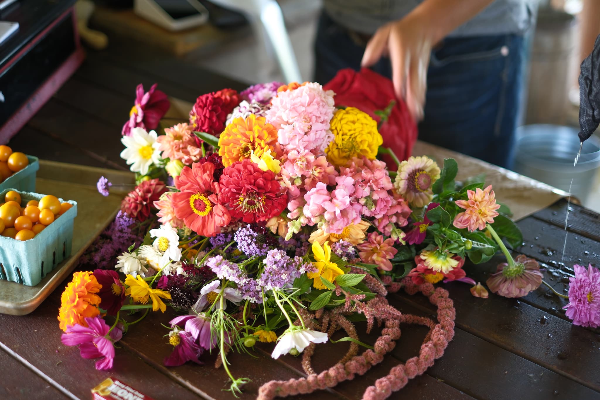 A colorful bouquet of various flowers on a wooden table, with a person arranging them in the background