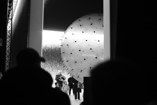 Silhouetted crowd watching performers on a stage with a large illuminated circular backdrop patterned with dots in a dark venue