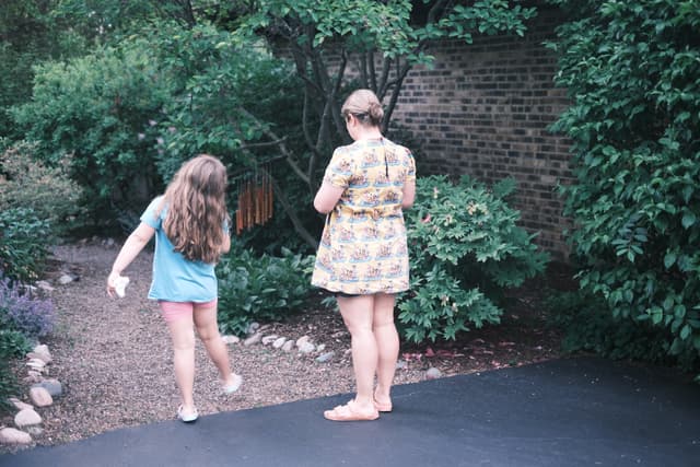 Two people standing outdoors near a garden with lush greenery and a brick wall in the background. One person is wearing a blue shirt and pink shorts, while the other is wearing a patterned dress