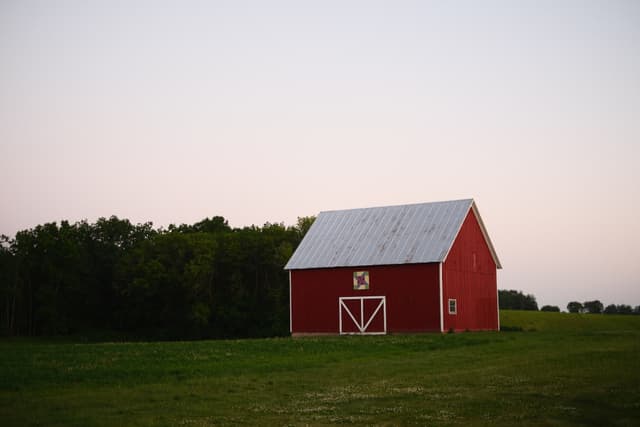 A red barn with a white roof stands in a grassy field, bordered by trees under a clear sky
