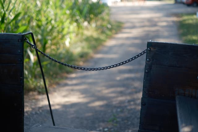 A dirt path bordered by greenery, viewed through an opening with a chain across it