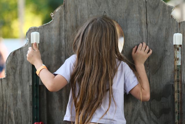A child with long hair peering through a wooden fence, gripping the posts on either side