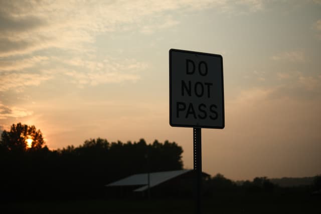 Silhouetted road sign reading DO NOT PASS against a warm sunset sky over a rural roadway