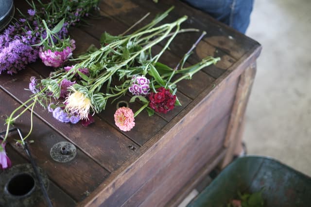 A wooden table with various cut flowers, including pink, purple, and red blooms, scattered on top. A green container is partially visible in the lower right corner