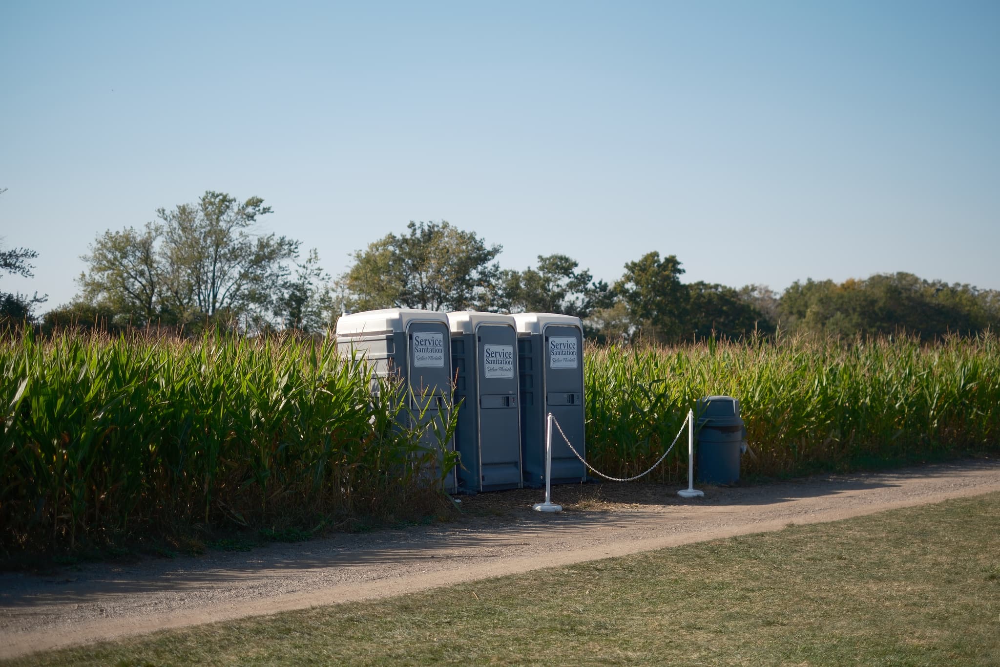 Two portable toilets are situated next to a cornfield, with a small trash bin nearby and a rope barrier in front