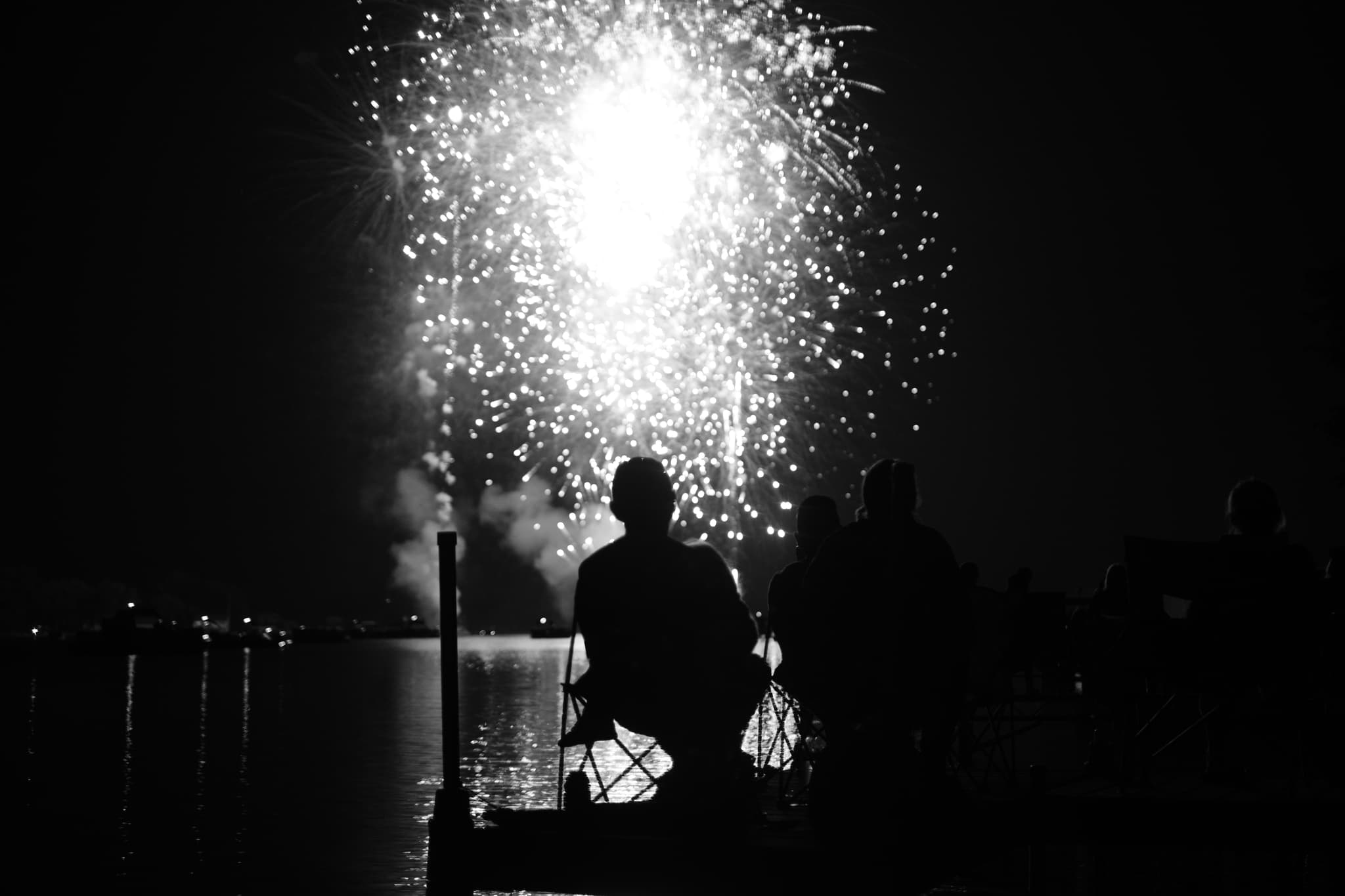 Silhouetted figures sitting on a dock, watching a bright fireworks display over a body of water at night