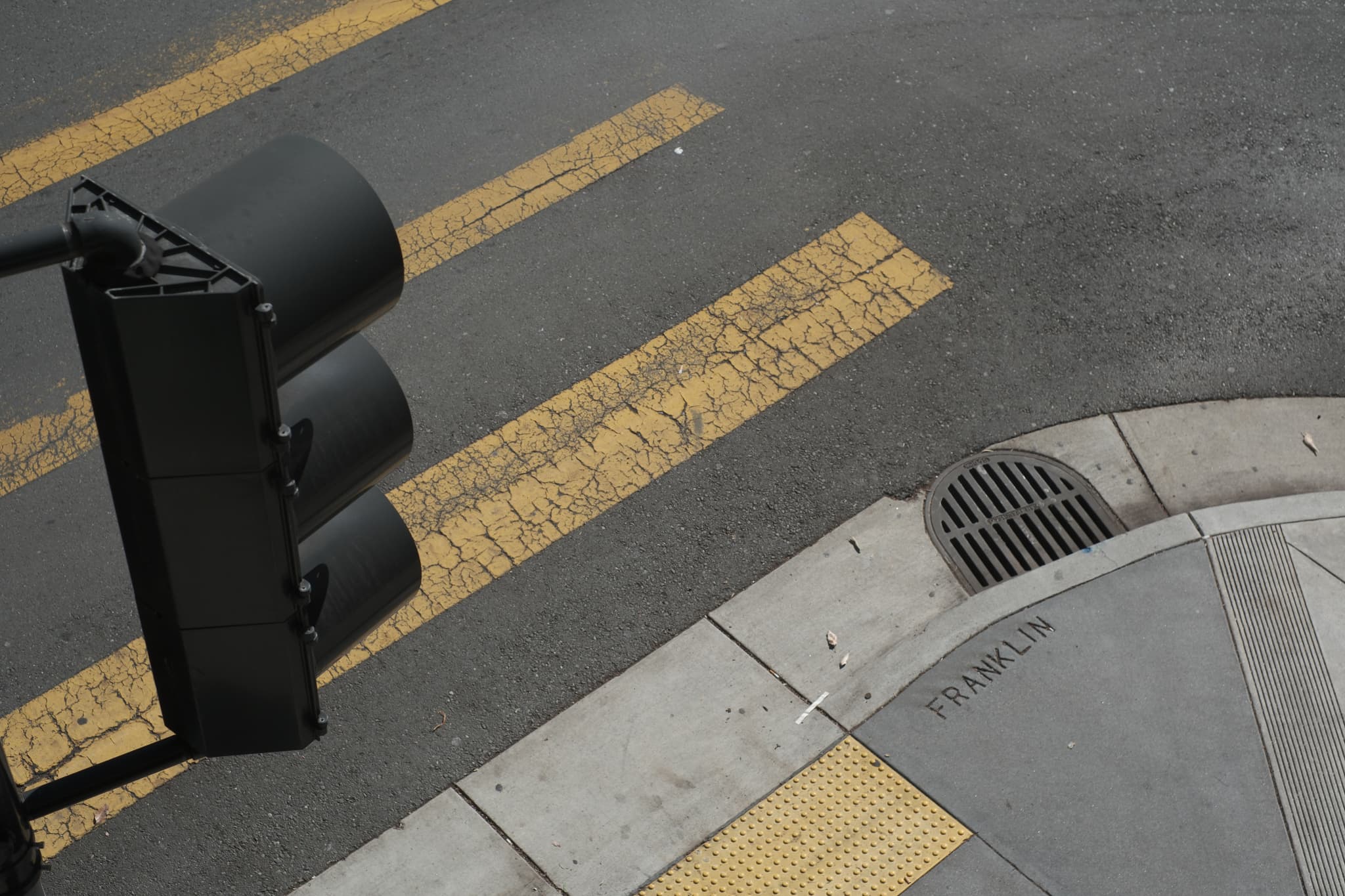 A traffic light at a street corner with yellow pedestrian crossing lines and a storm drain on the sidewalk