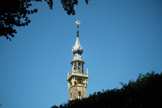 A tall, ornate tower with a spire and clock, set against a clear blue sky, partially framed by silhouetted tree branches