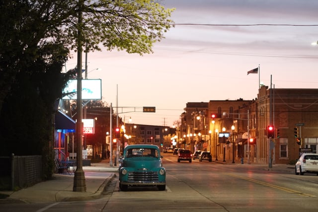 A quiet street scene at dusk with a vintage car parked on the side, illuminated by streetlights and surrounded by buildings