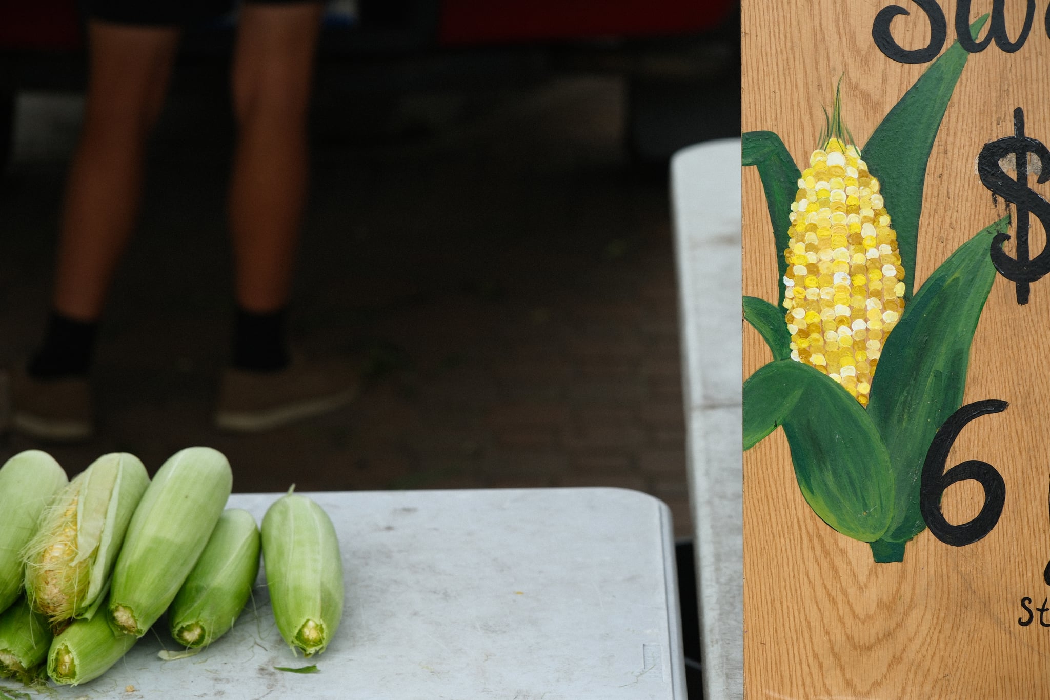 Close-up of several ears of corn on a table beside a hand-painted sign with a corn illustration and a price at a market stand