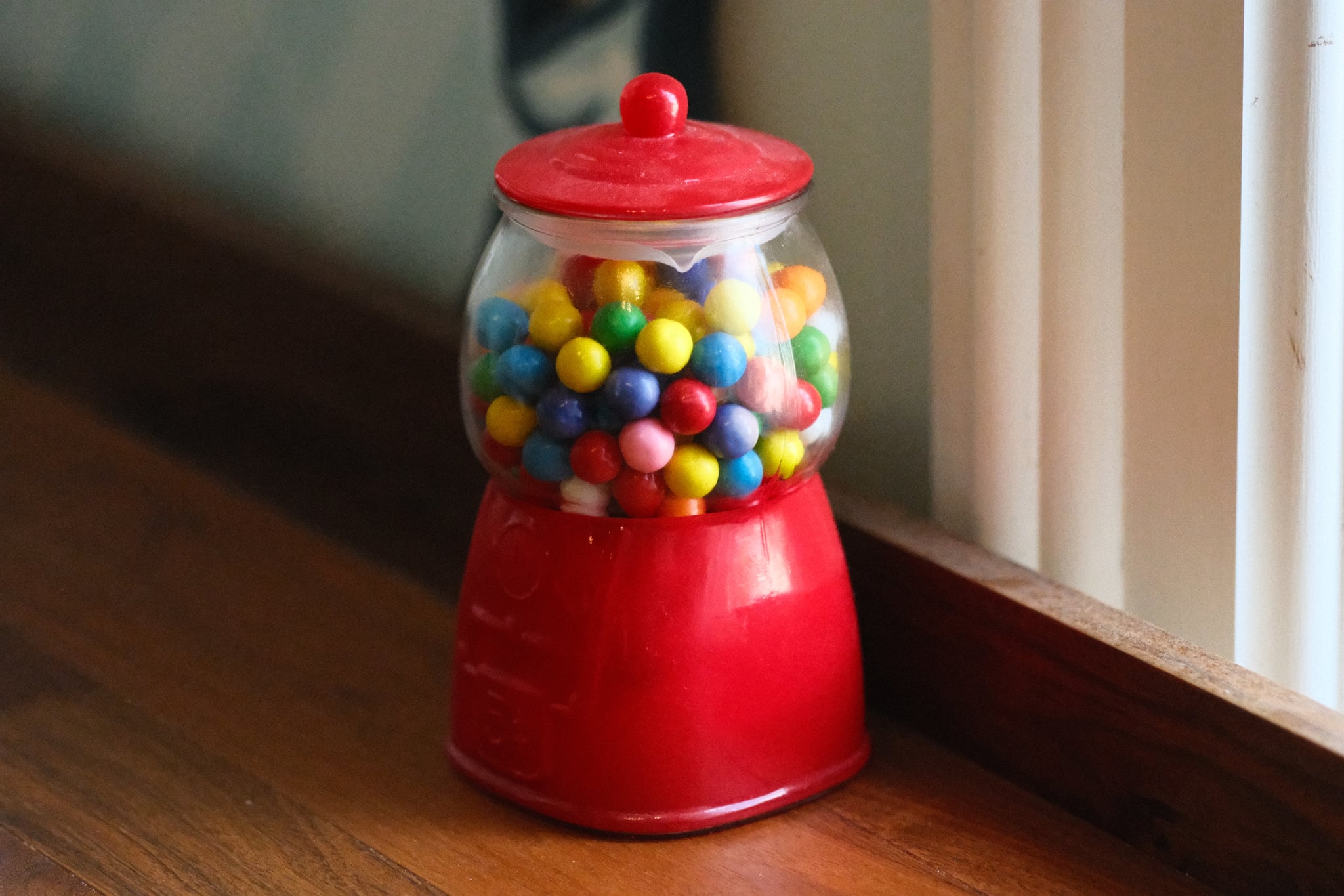 A red gumball machine filled with colorful gumballs, placed on a wooden surface near a window