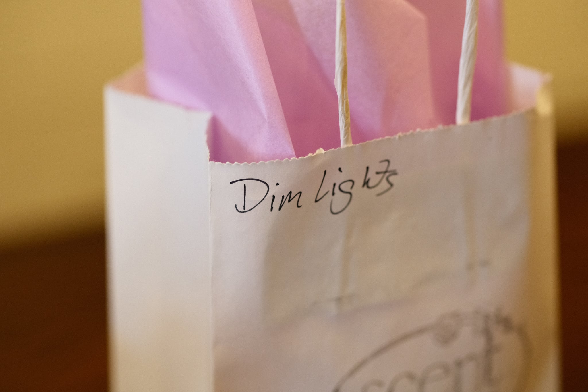 Close-up of a white gift bag with pink tissue paper and handwritten text reading “Dim light” near the top, set against a warm indoor background
