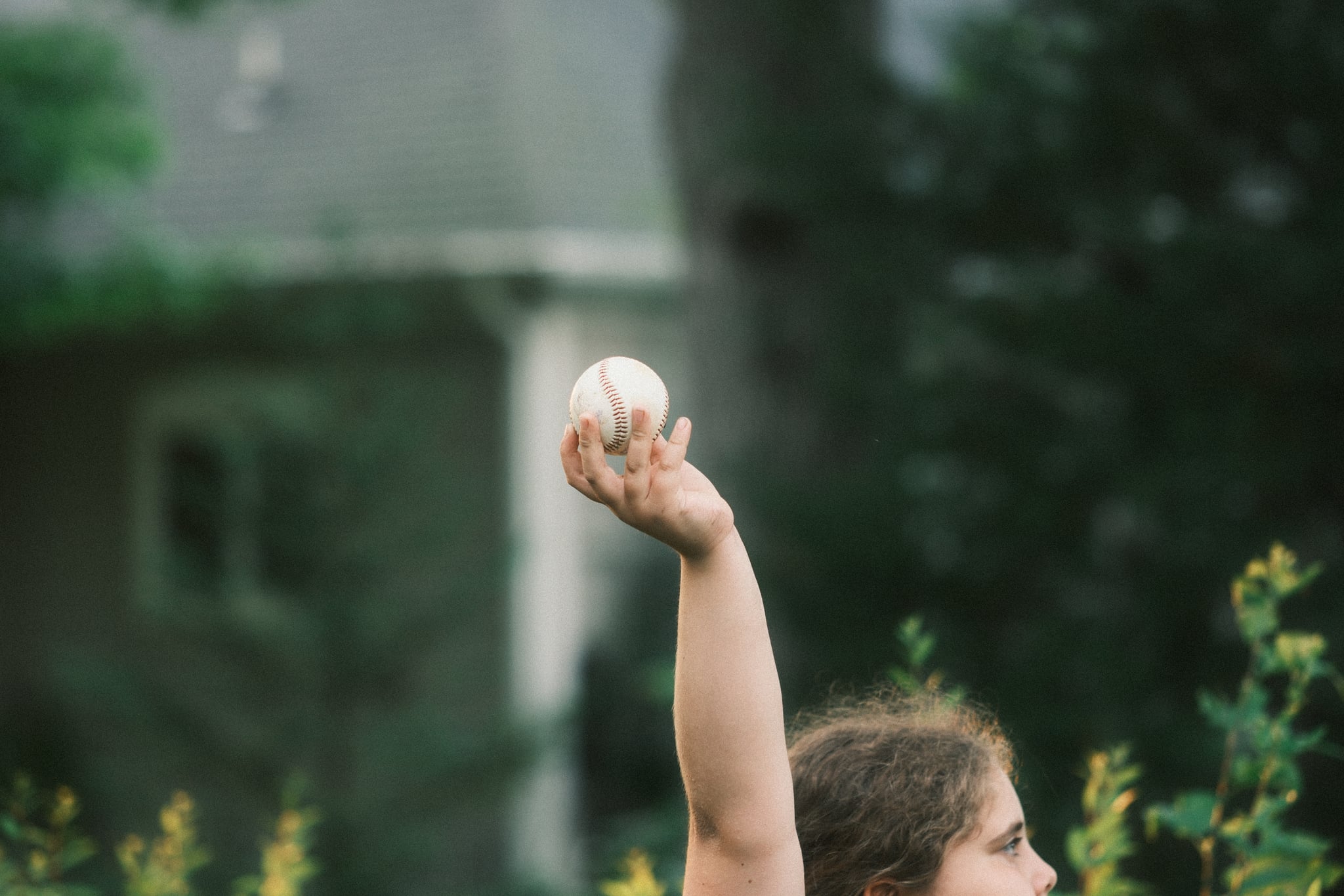 Person outdoors with arm raised holding a small object, with a house and trees blurred in the background