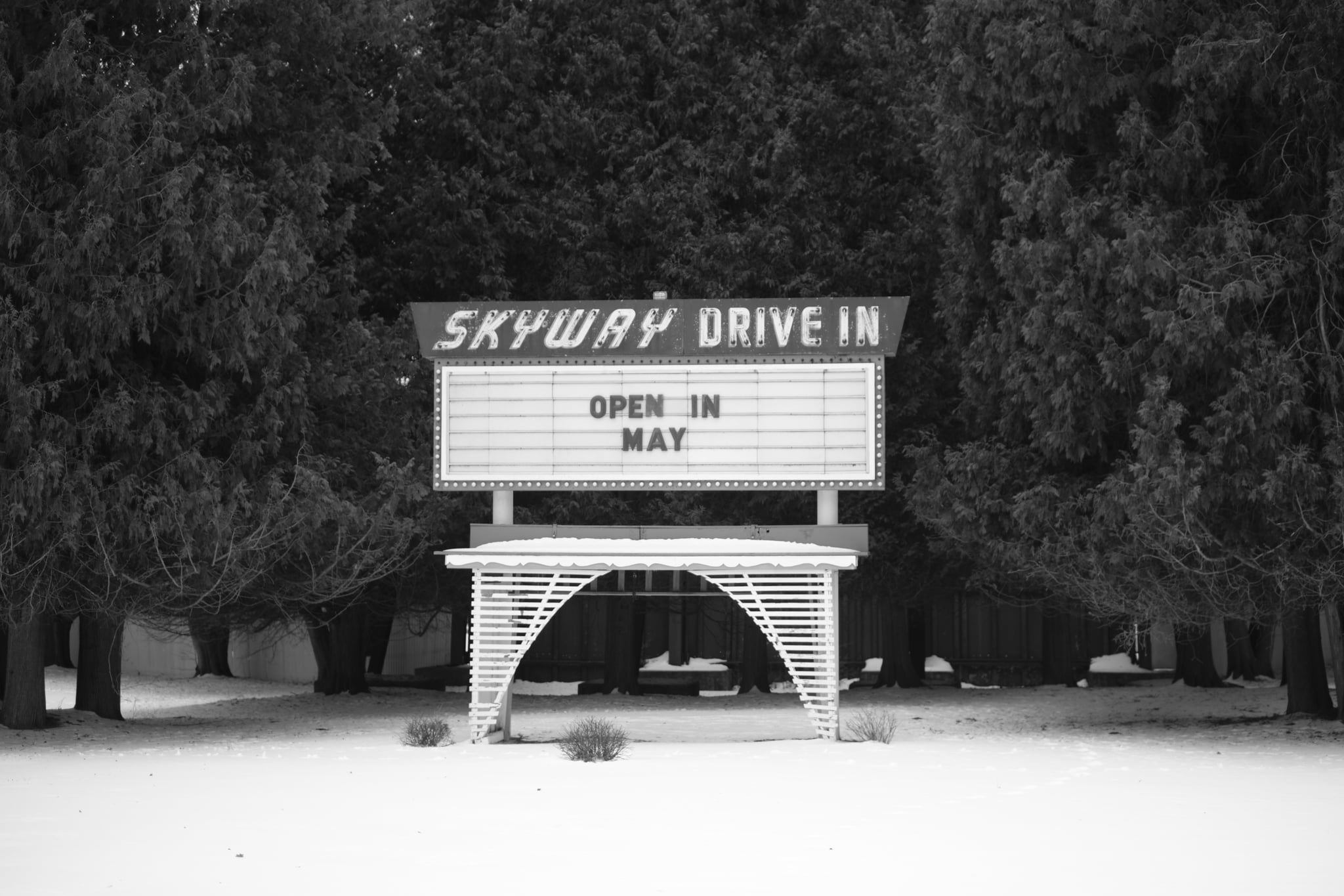 A black and white photo of a Skyway Drive In sign surrounded by trees, with the message Open in May