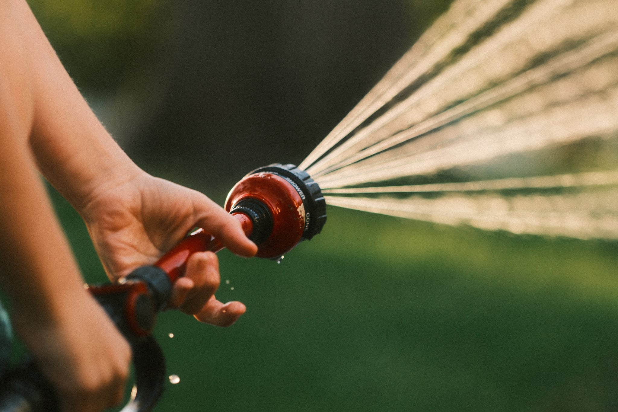 A hand holding a garden hose nozzle spraying water with a blurred green background