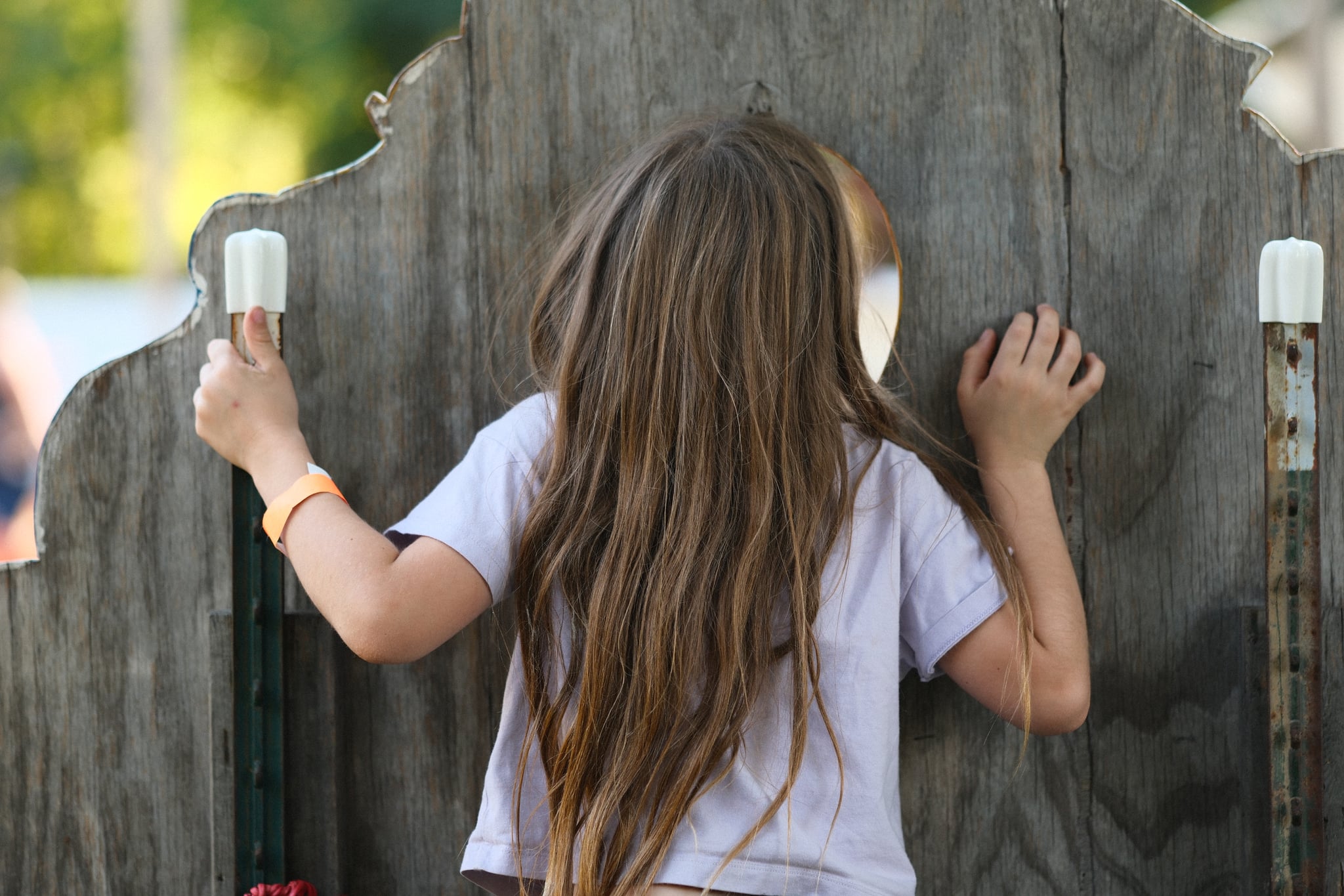A child with long hair peering through a wooden fence, gripping the posts on either side