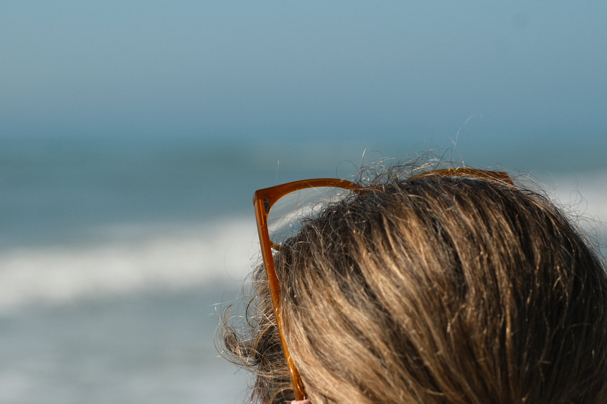 Close-up of a person with light brown hair and sunglasses on their head, facing the ocean with waves in the background