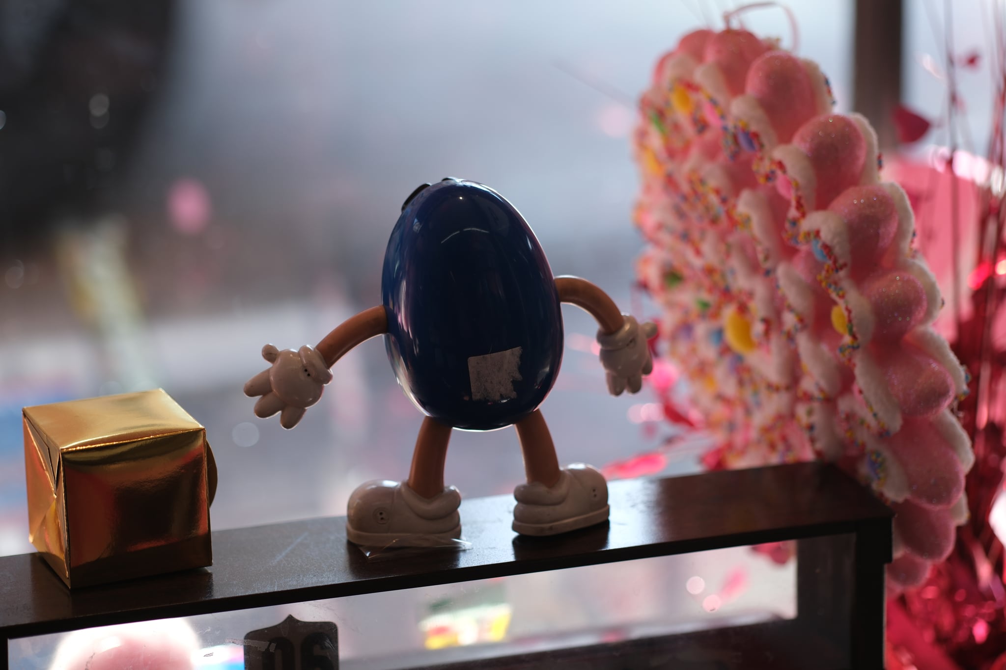 Toy figure with dark hair leaning forward on a glass table beside a small gold box and a pink flower arrangement, with a blurred cherry-blossom background