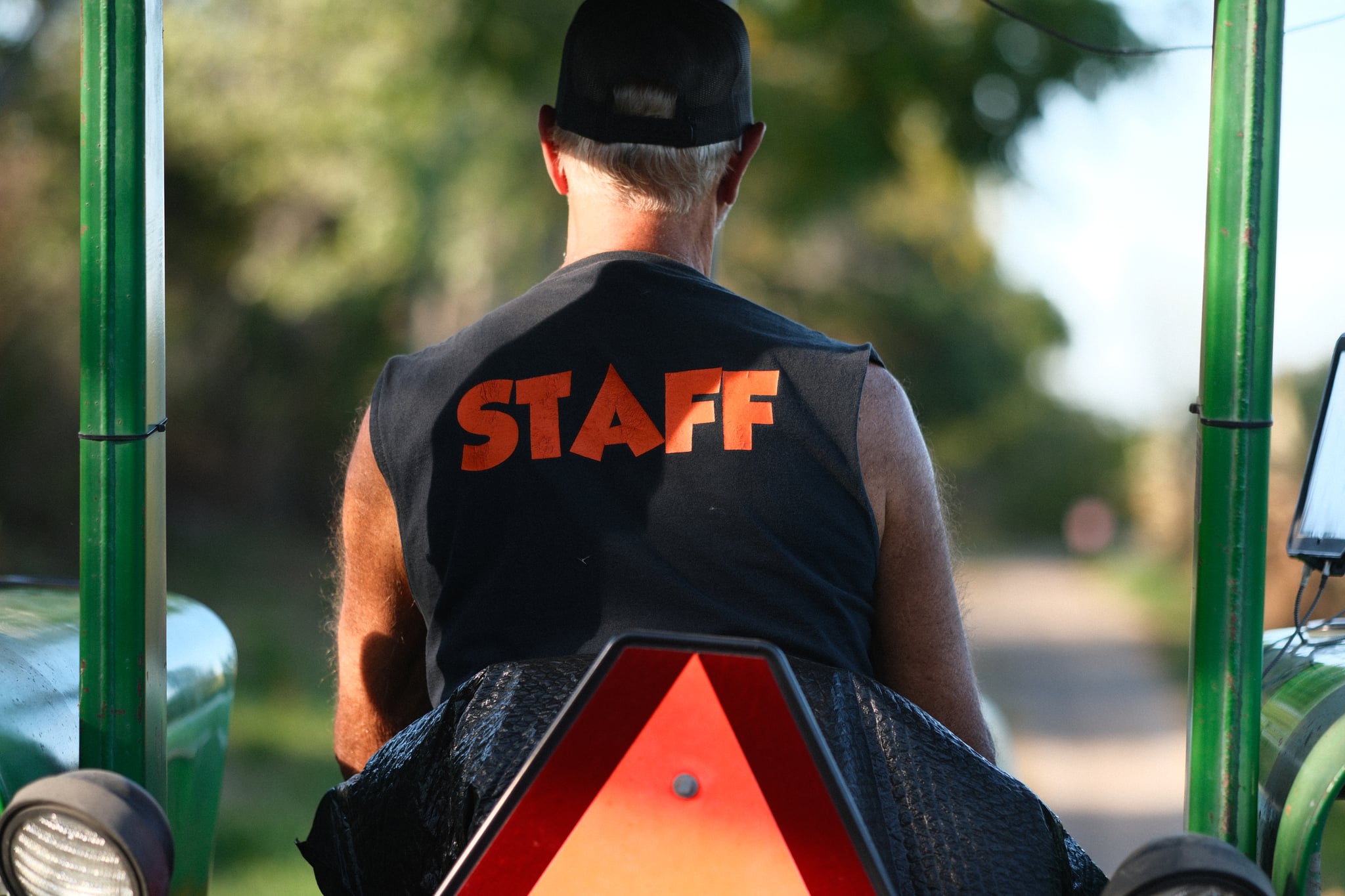 A person wearing a 'STAFF' vest and a black cap, sitting on a vehicle with a triangular reflective sign, surrounded by green outdoor scenery