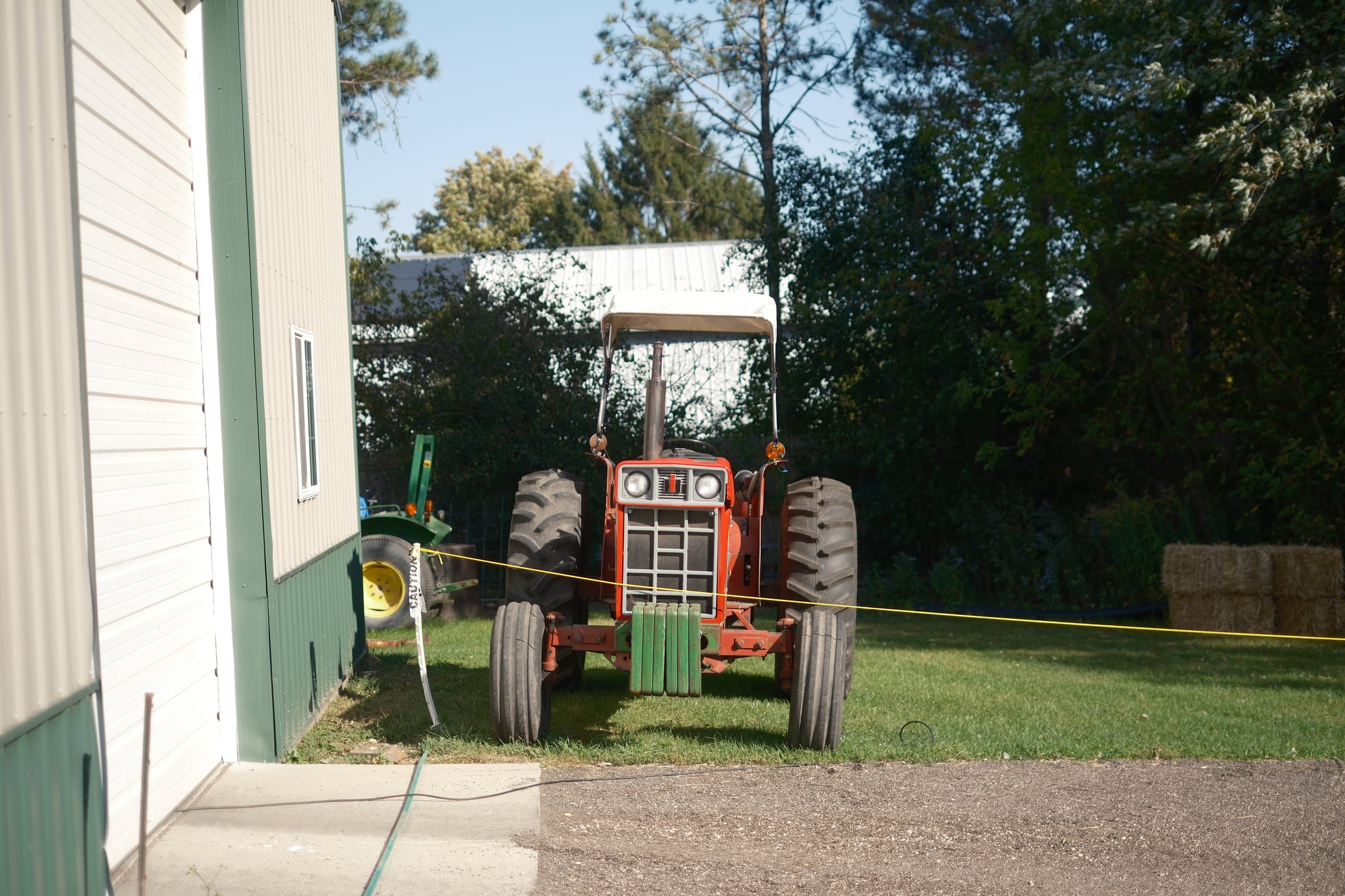 A red tractor parked on grass near a building, with trees and hay bales in the background