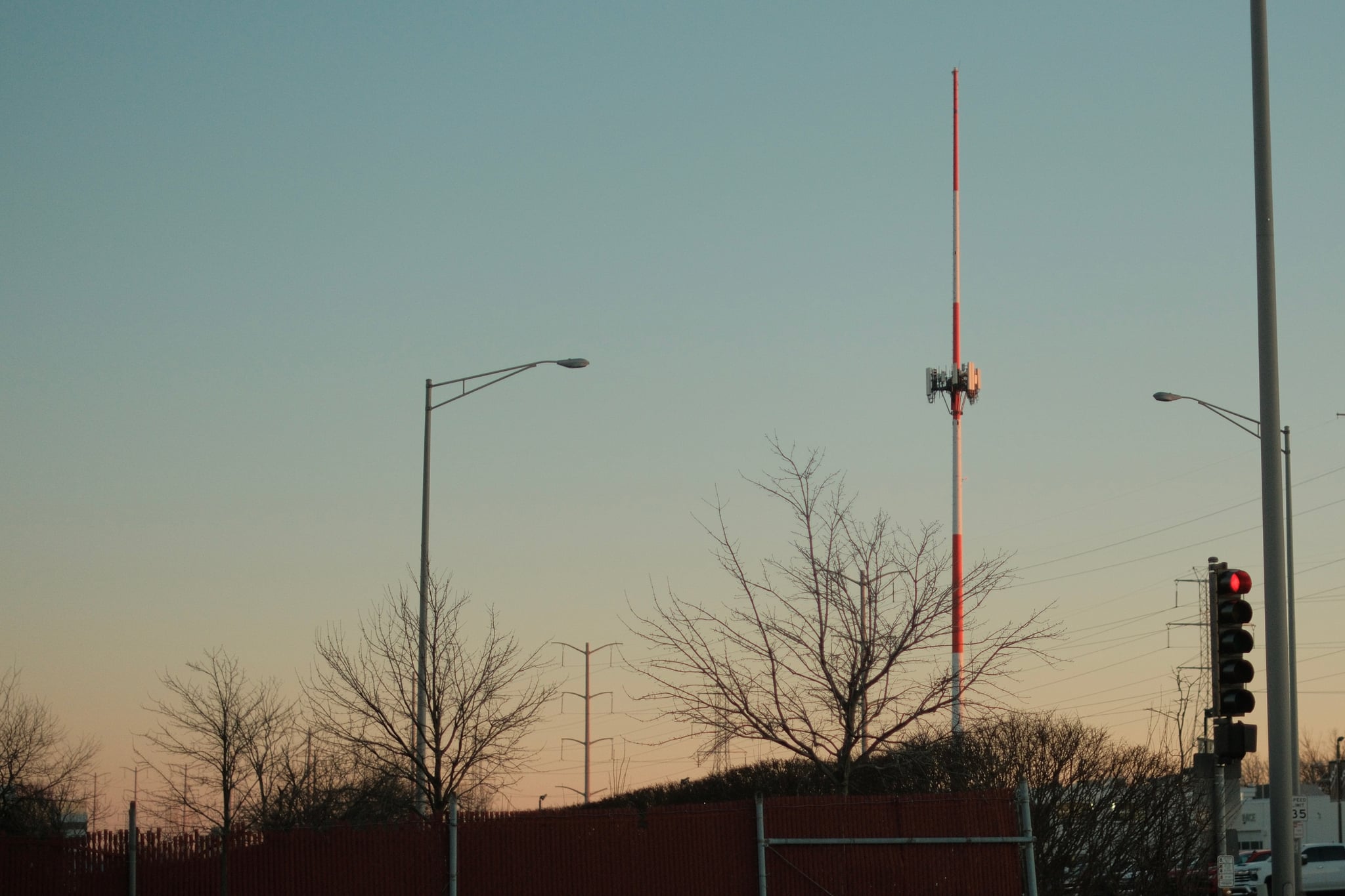 A tall radio tower with a red and white antenna stands against a clear sky, surrounded by bare trees and streetlights. A traffic light is visible in the foreground