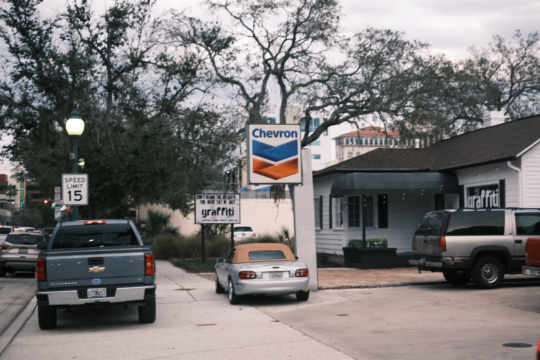 A street scene with parked cars, a Chevron gas station sign, and houses in the background