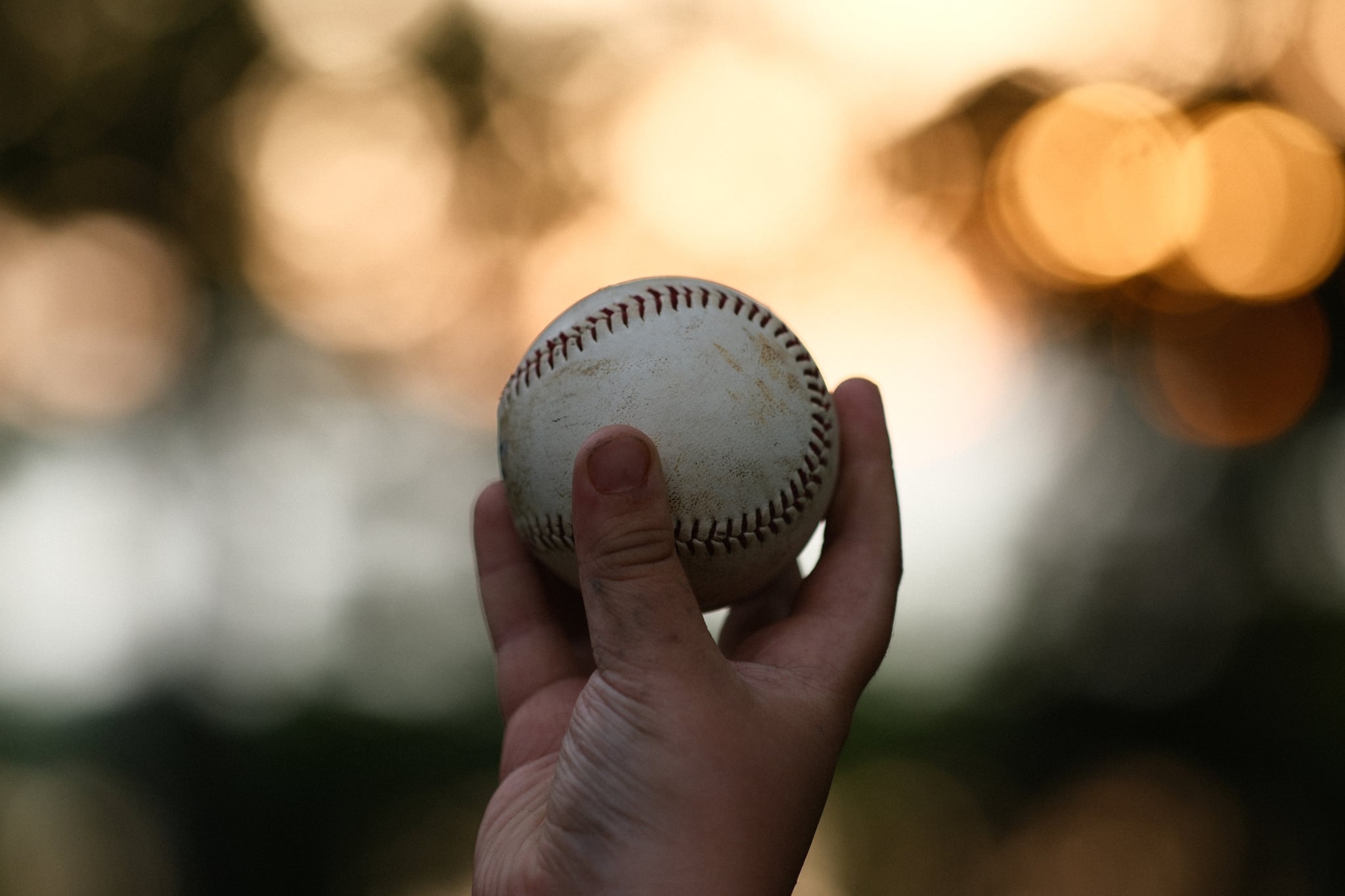 Hand holding a baseball up against a softly blurred background with warm bokeh lights, suggesting an outdoor setting near sunset