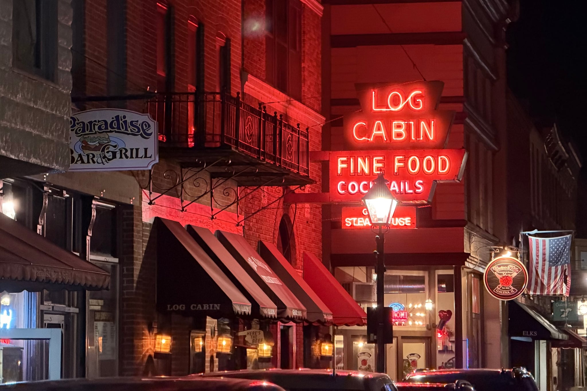 Red neon “Log Cabin Fine Food Cocktails” sign glowing above a cozy street-side restaurant at night