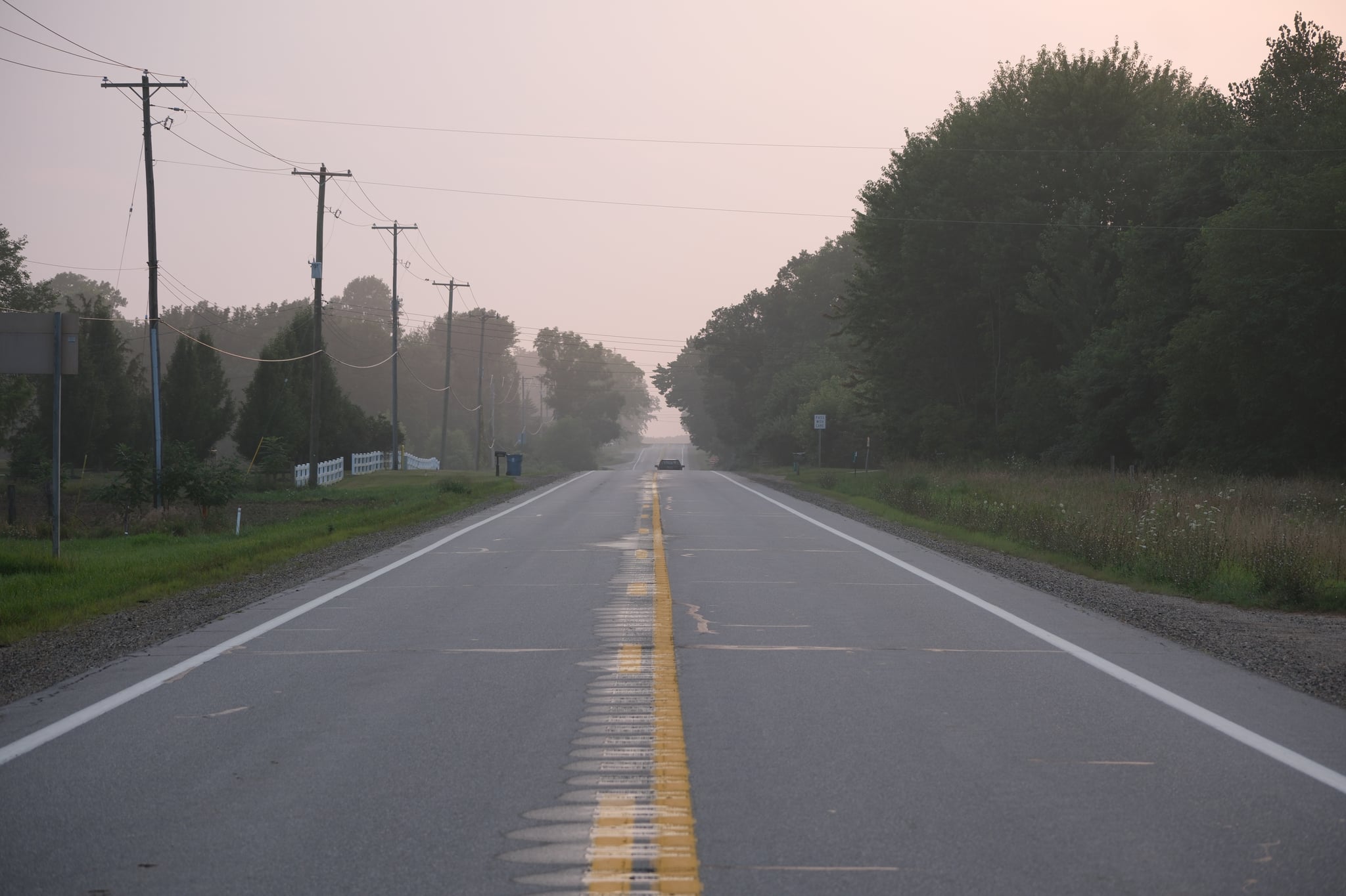 Empty two-lane highway stretching into the distance at dawn, lined with trees and utility poles under a hazy sky