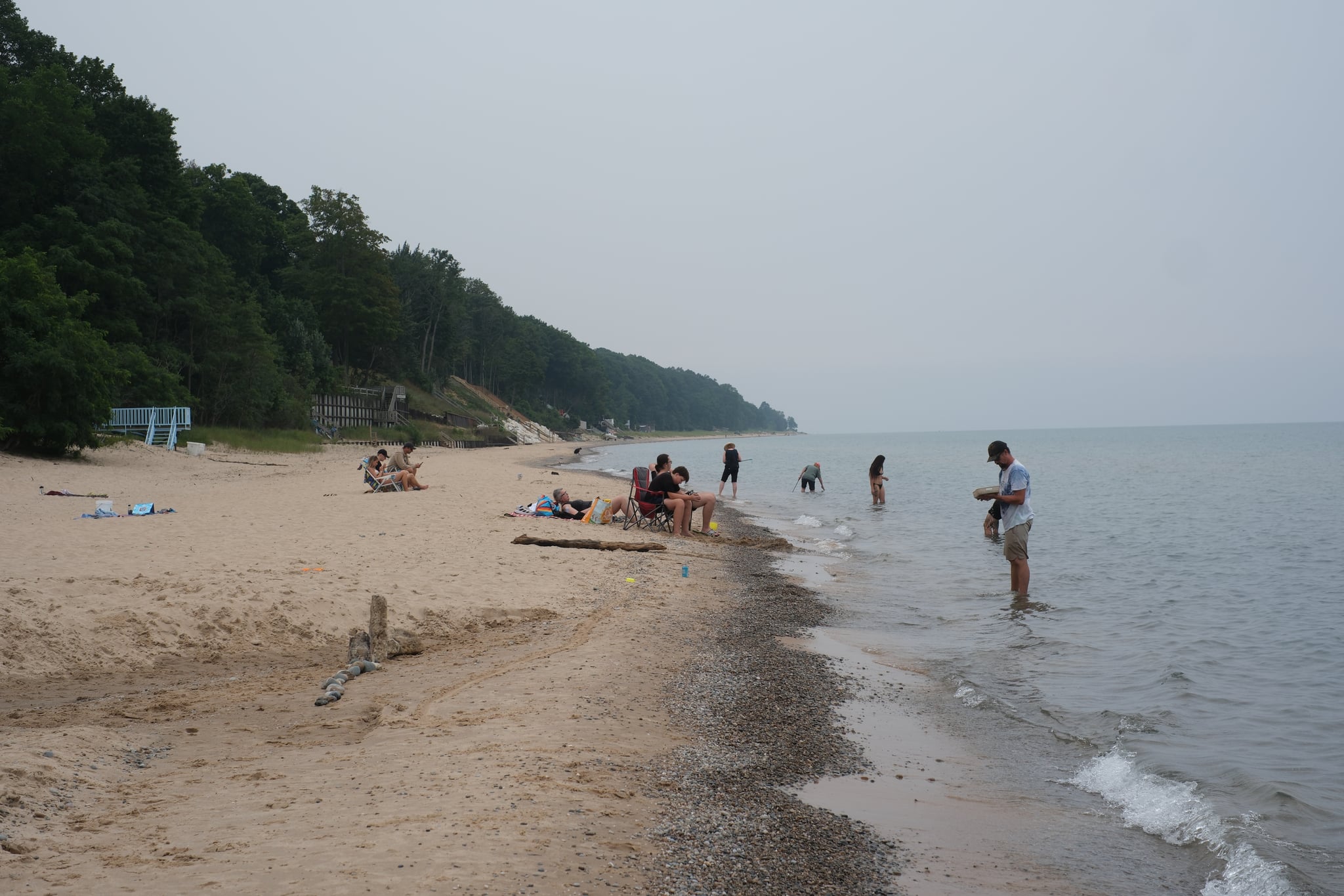 Hazy shoreline with scattered beachgoers wading and relaxing near gentle waves along a tree-lined coast