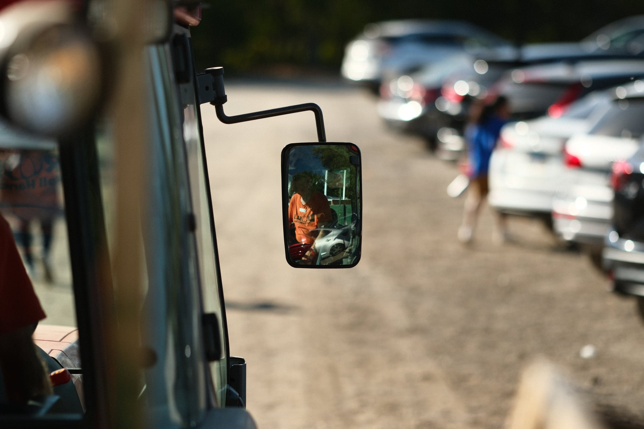 Rearview mirror reflecting a smiling woman's face, with parked cars and a blurred figure in the background