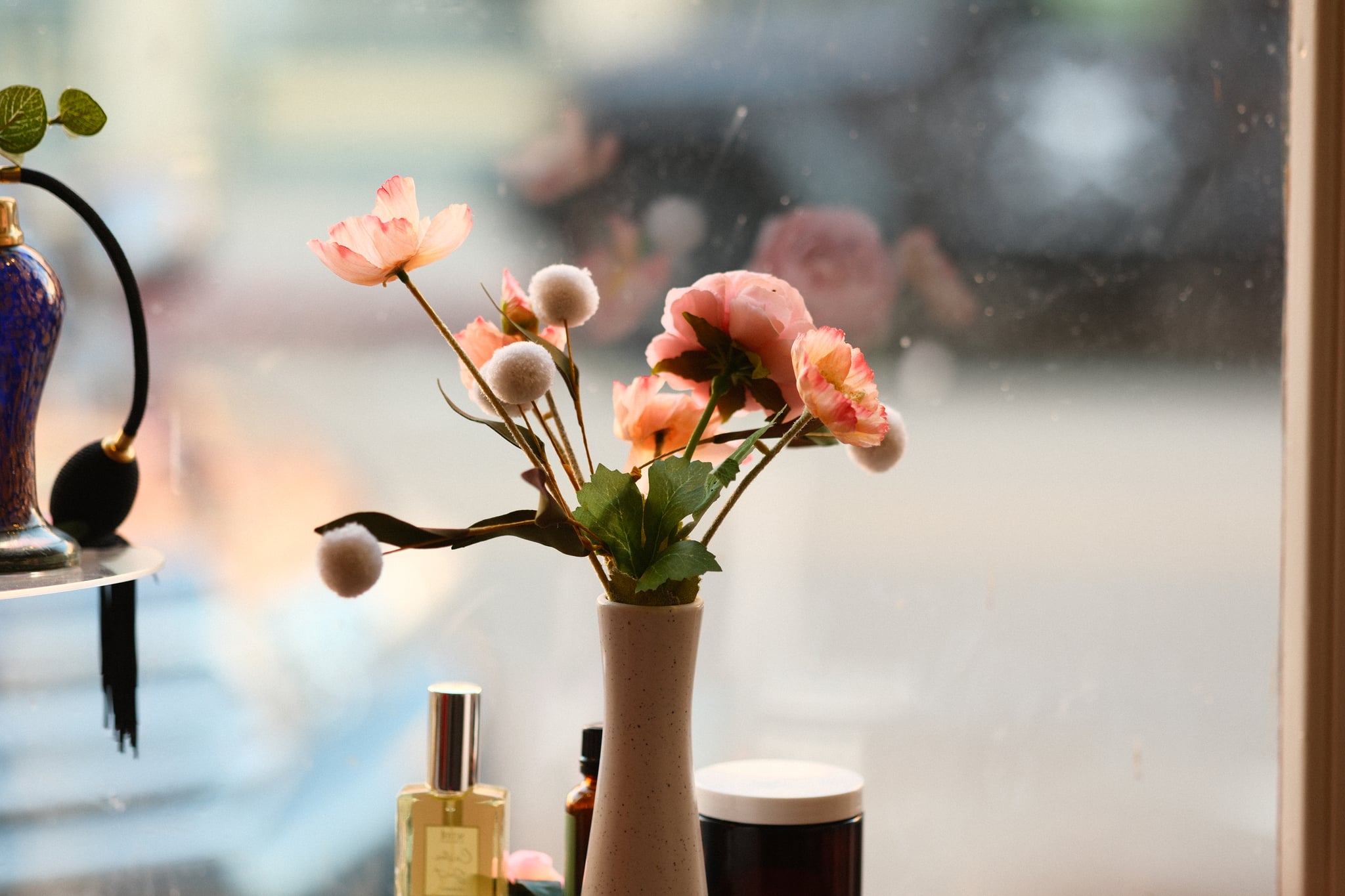 Soft-focus window-side still life with a vase of pink and white flowers, small cosmetics or perfume bottles on a table, and warm natural light with a blurred city street outside