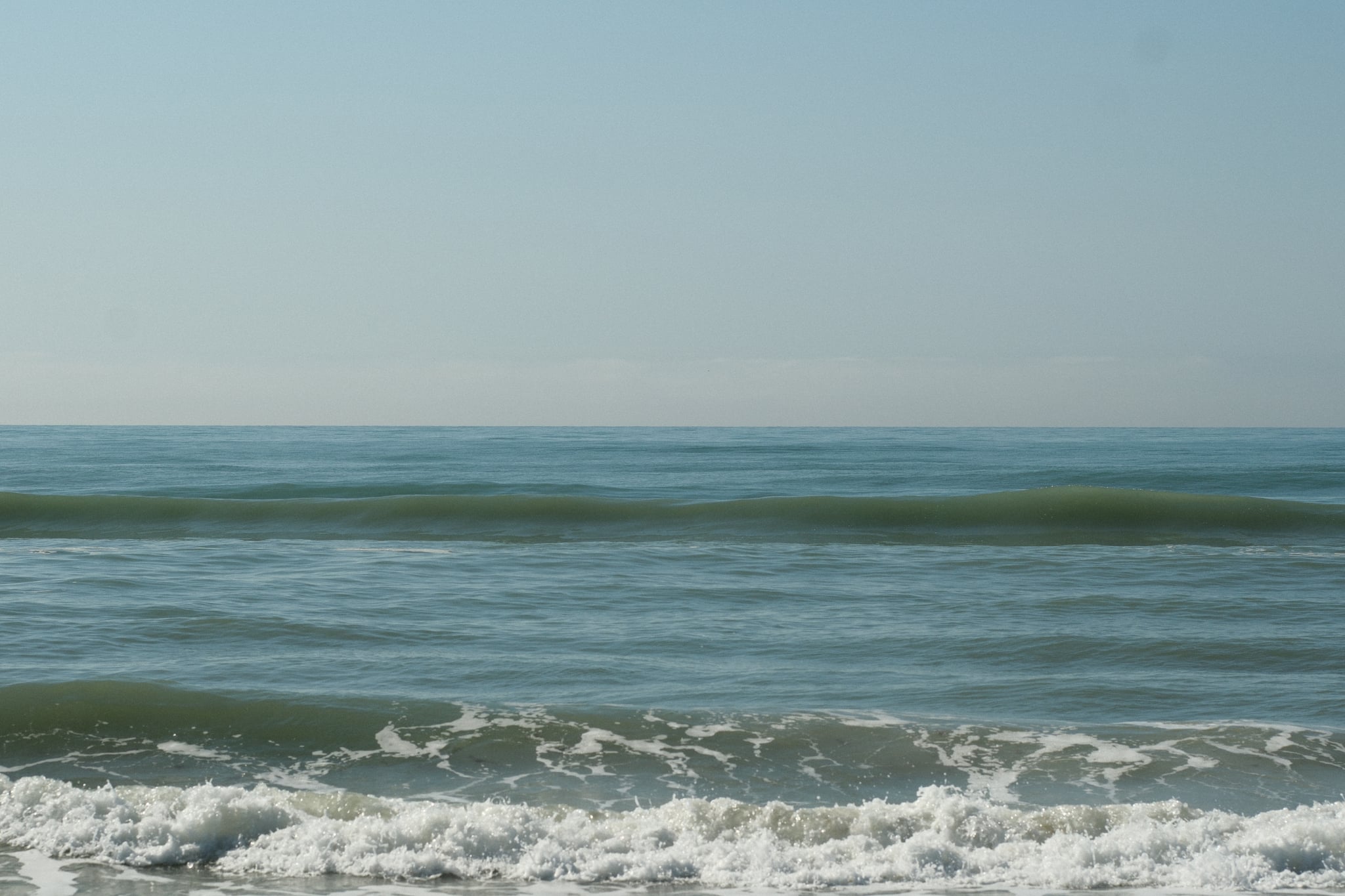 Small ocean waves rolling toward a sandy shoreline under a clear sky, with a low horizon across the calm sea