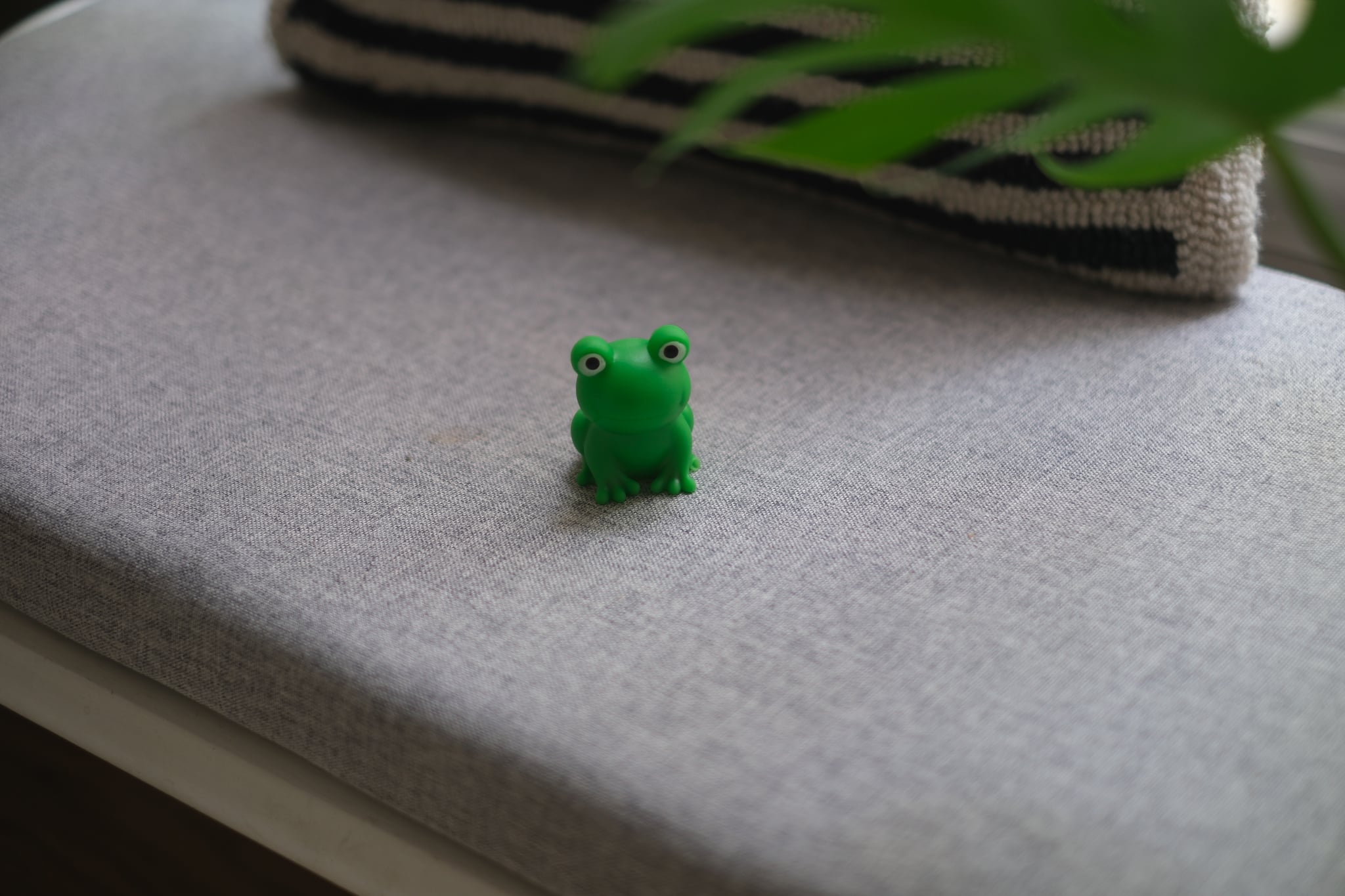 A small green frog figurine placed on a gray cushioned surface with a striped pillow and a green plant in the background