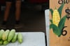 Close-up of several ears of corn on a table beside a hand-painted sign with a corn illustration and a price at a market stand