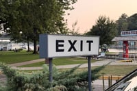 Large roadside EXIT sign on metal posts near a parking area and trees at dusk