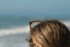 Close-up of a person with light brown hair and sunglasses on their head, facing the ocean with waves in the background