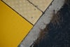 A close-up of a sidewalk corner with yellow tactile paving, adjacent to a concrete curb and asphalt road, with some grass growing in the cracks