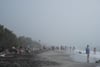 A misty beach scene with people scattered along the shore, some in the water, and others lounging or walking on the gray sand