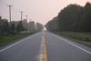 Empty two-lane highway stretching into the distance at dawn, lined with trees and utility poles under a hazy sky