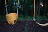 A yellow toddler swing hangs from metal chains against a blurred background of trees and foliage, suggesting a peaceful outdoor setting, possibly a park or a backyard