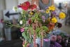 A vibrant bouquet of various flowers in a metal bucket, featuring red, yellow, purple, and white blossoms, with a person in the background